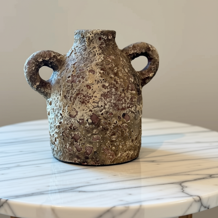 A handmade stone vase with a textured, speckled finish in earthy brown, beige, and gray tones, featuring two loop handles, displayed on a white marble-topped table against a neutral beige wall.