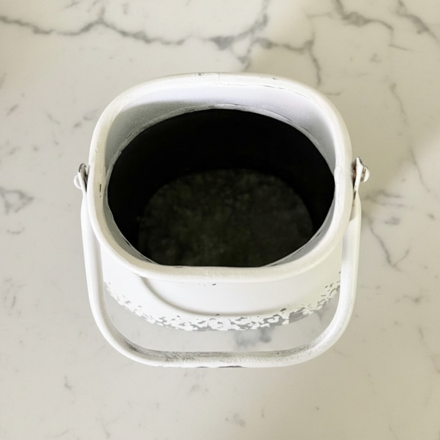 A white metal vintage milk can viewed from above, showing a dark interior and a decorative silver pattern around the lower rim, placed on a white marble surface.