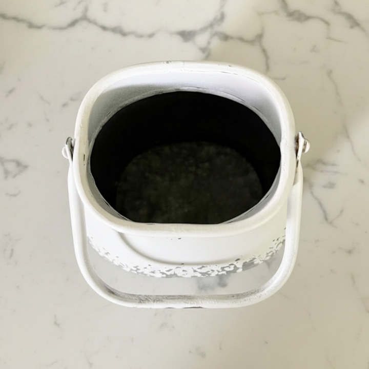 A white metal vintage milk can viewed from above, showing a dark interior and a decorative silver pattern around the lower rim, placed on a white marble surface.