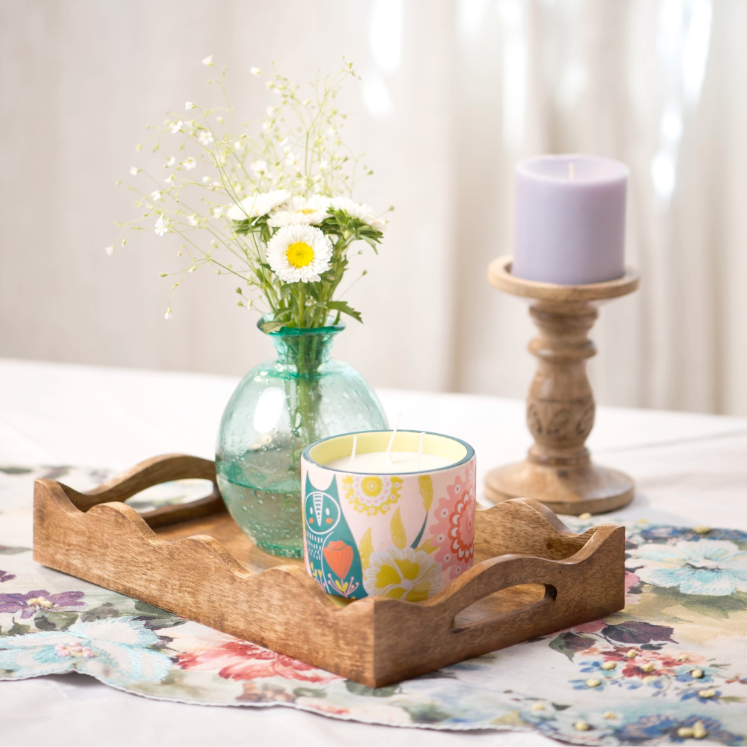 Wooden tray with a vase of flowers, colorful mug, and candle on a floral tablecloth.