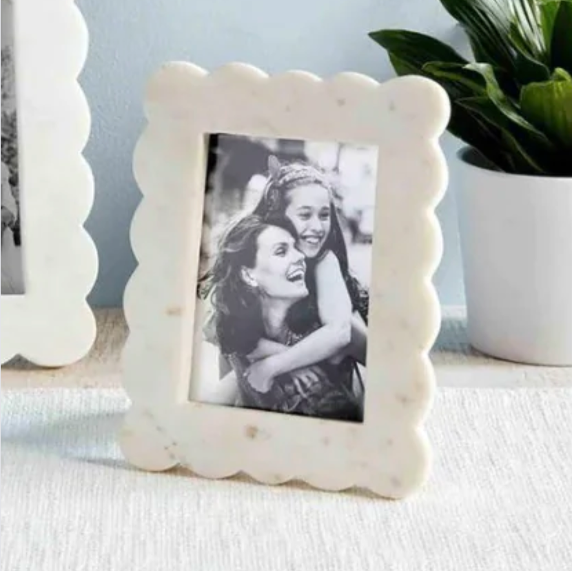 A white marble picture frame with scalloped edge details, holding a photo of two people, placed on a surface with a plant in the background.