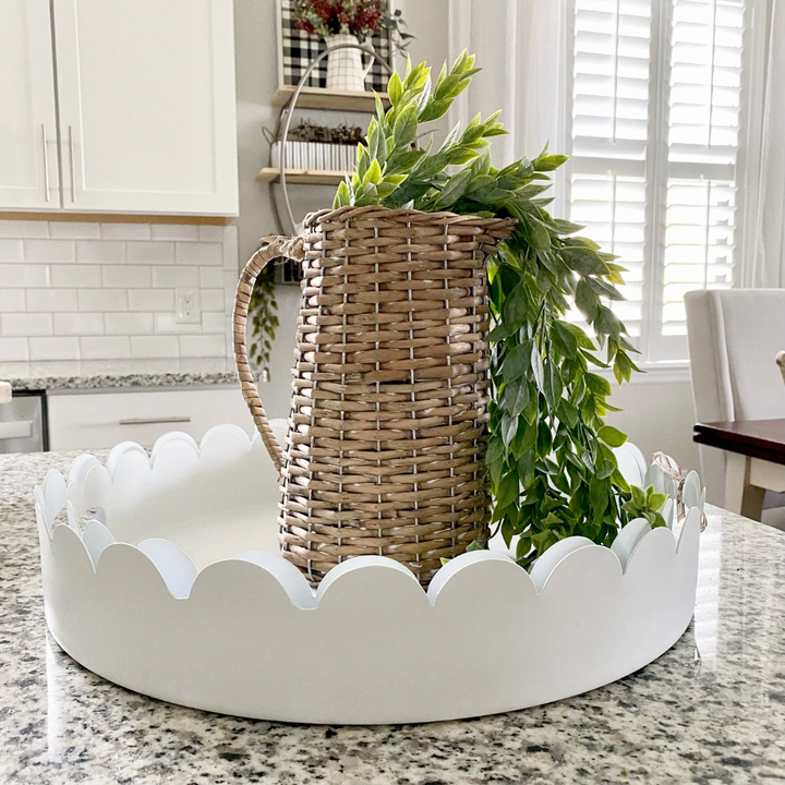 White scalloped-edge tray with a wicker pitcher and greenery on a kitchen counter.