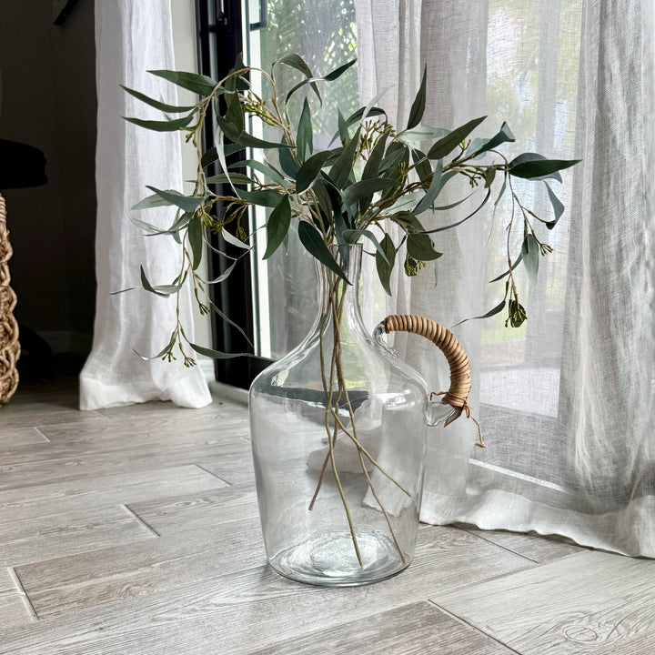 Clear glass vase with seeded eucalyptus stems on a gray wooden floor with curtains in the background