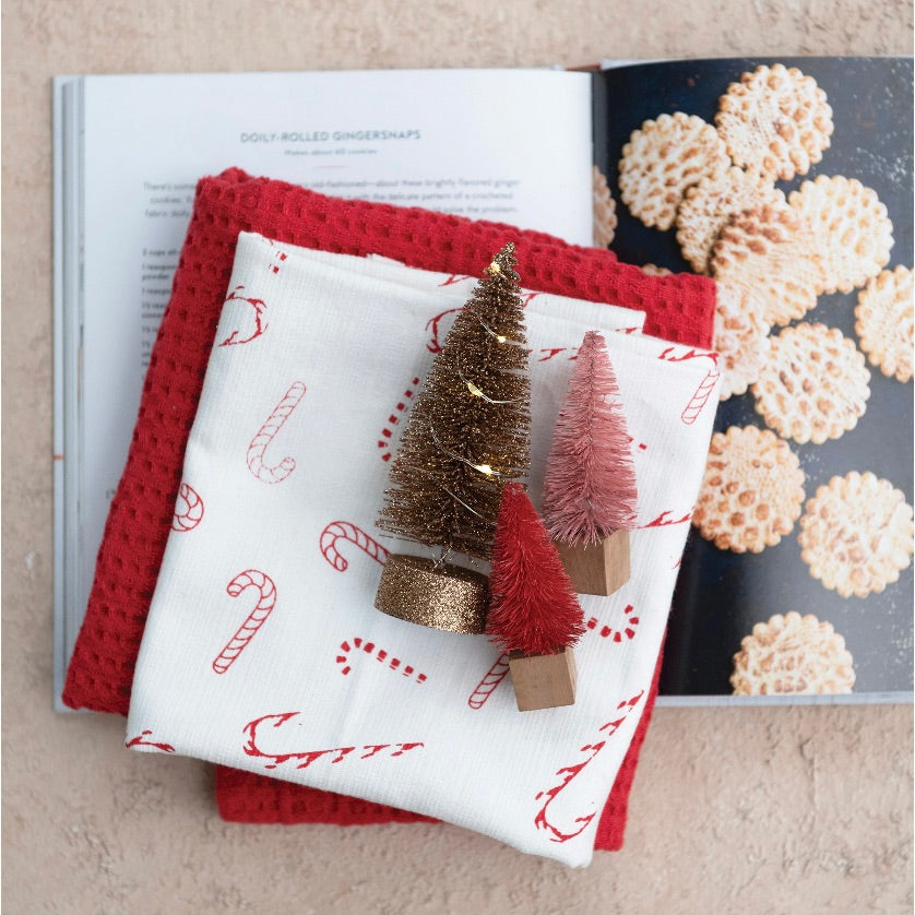 Two folded kitchen towels with a white candy cane pattern on a red background and a solid red waffle weave textured towel, displayed on a flat surface.