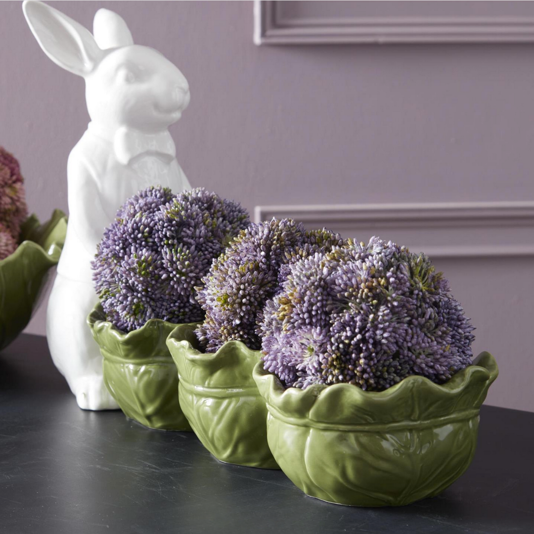 Three green cabbage-shaped planters with purple sedum balls on a dark surface, with a white ceramic rabbit figure in the background.
