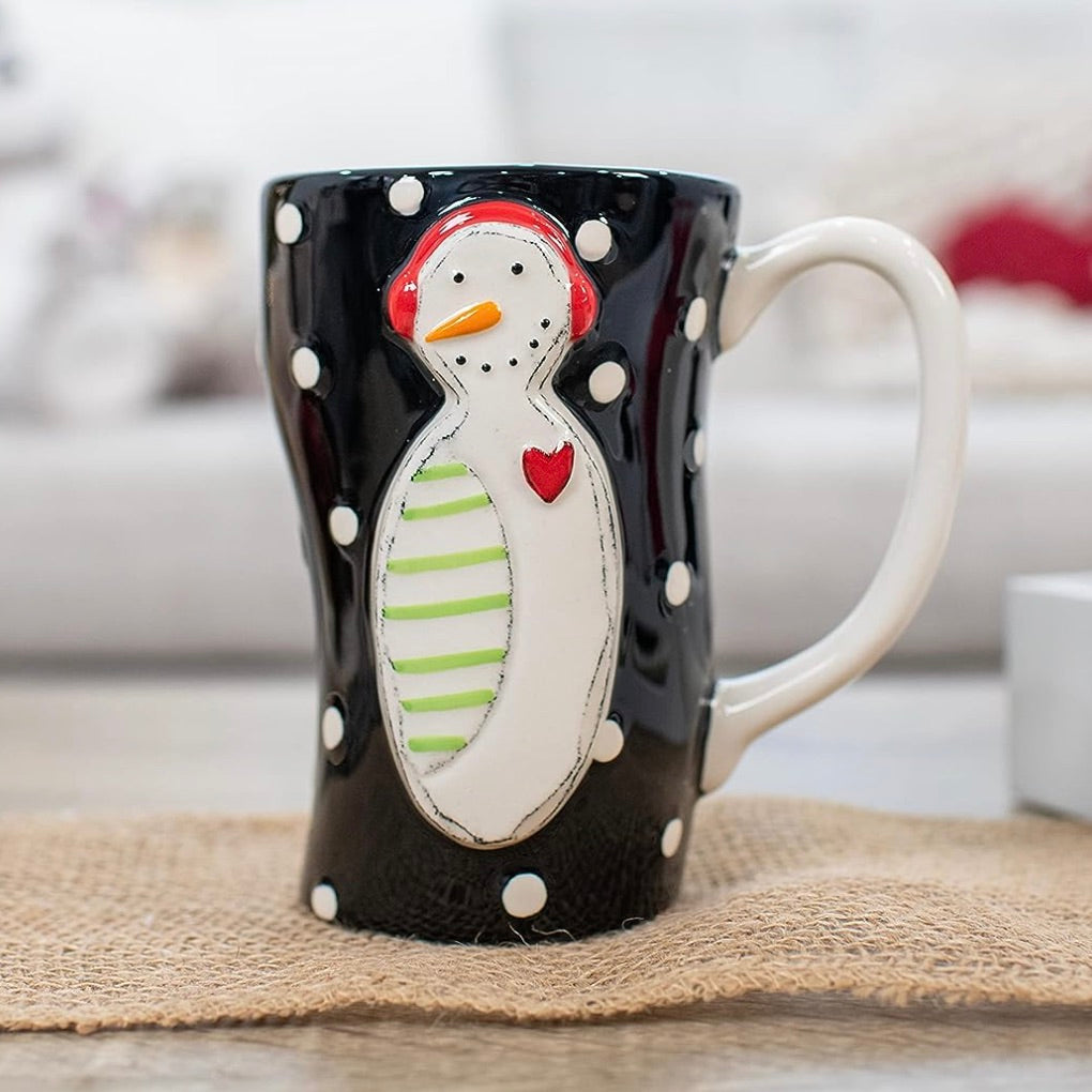 A ceramic mug with a black and white polka dot pattern featuring a stylized snowman character with a red nose, green scarf, and white earmuffs.