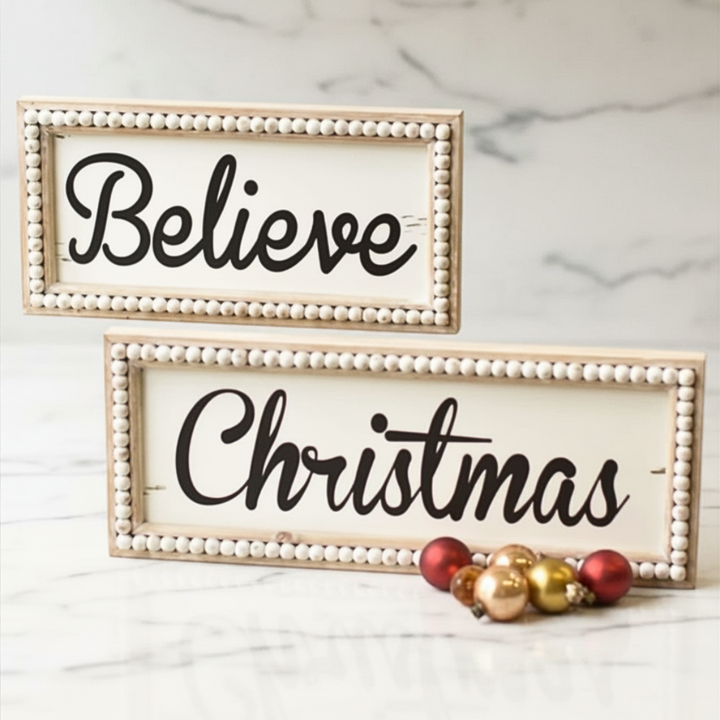 Two wooden beaded Christmas signs with the words 'BELIEVE' and 'CHRISTMAS' written in black script, placed on a rustic wooden surface with pine needles and holiday ornaments around.