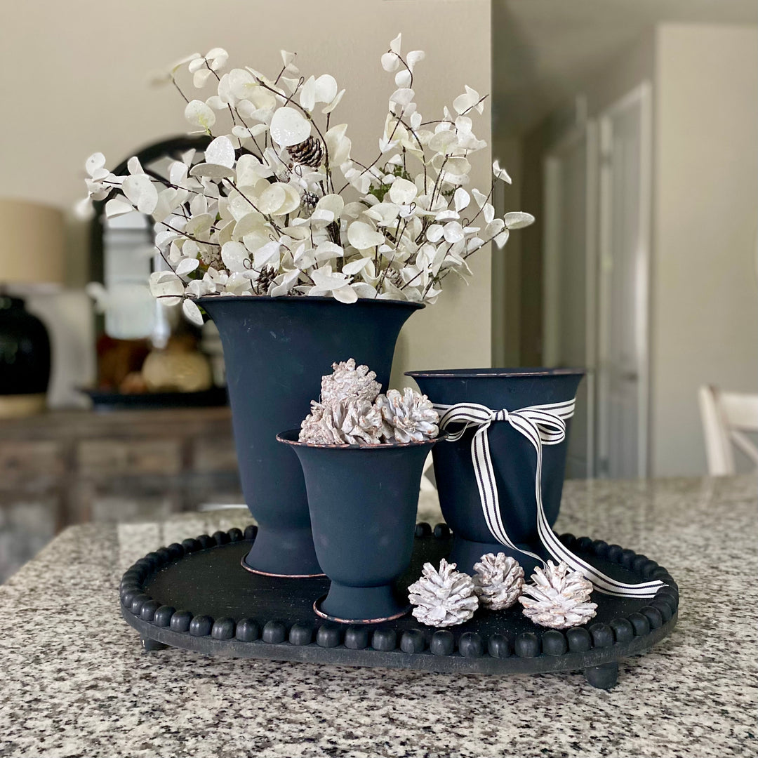 A set of three black metal urns of varying sizes arranged on a surface, with white floral arrangements inside the larger two urns and decorative items like pine cones. The urns have a matte finish and a rustic look.
