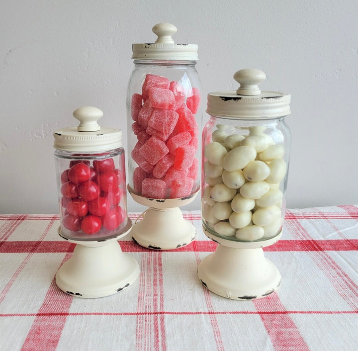 Three distressed apothecary glass canisters with lids, two containing red and white candy, and one with white and red candy. Placed on a table with a red and white checkered tablecloth.