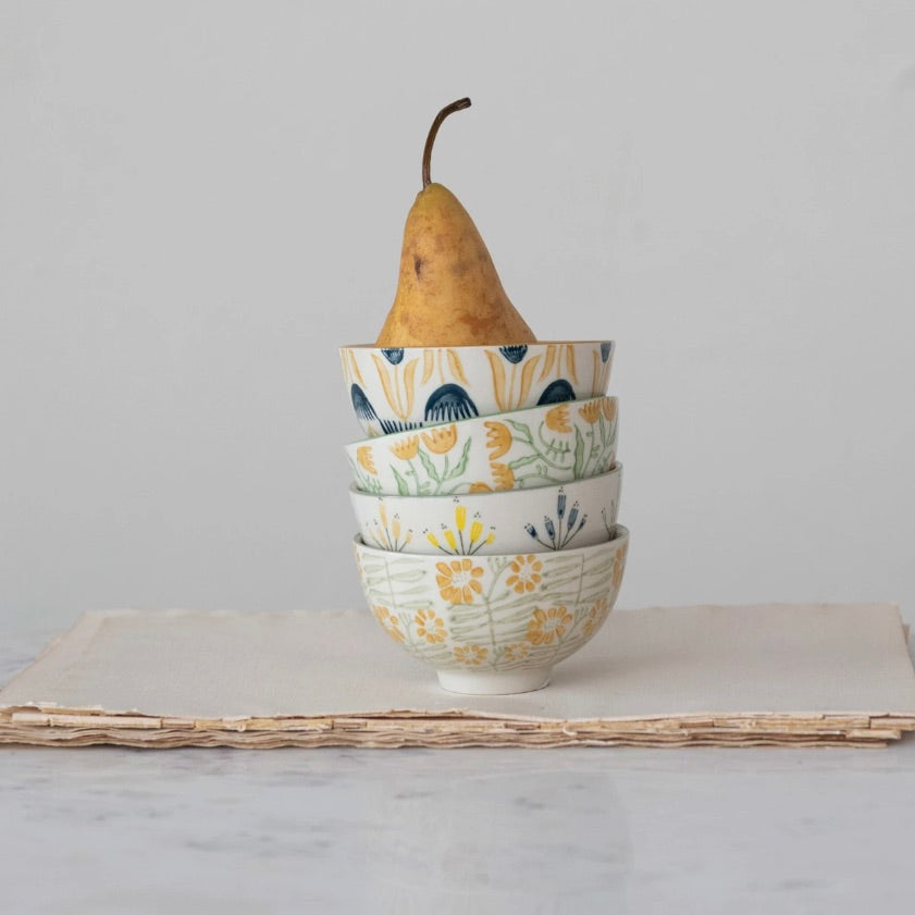 A set of four stoneware bowls with floral patterns in yellow and white colors, stacked on top of each other with a pear on the top bowl, placed on a paper-like surface.