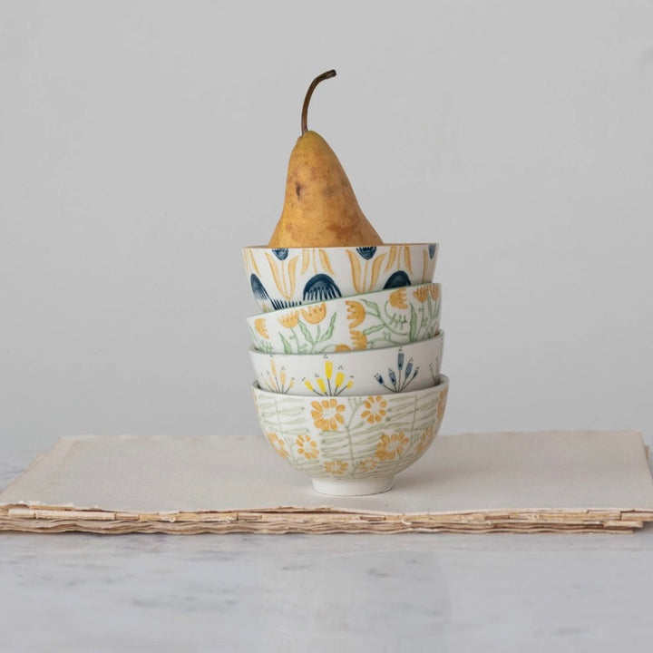 A set of four stoneware bowls with floral patterns in yellow and white colors, stacked on top of each other with a pear on the top bowl, placed on a paper-like surface.