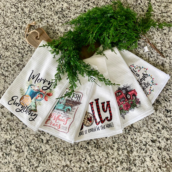A set of white Christmas kitchen towels with various festive designs and text such as 'Merry Christmas' and 'Jolly', displayed on a surface with a green decorative branch and a tag.