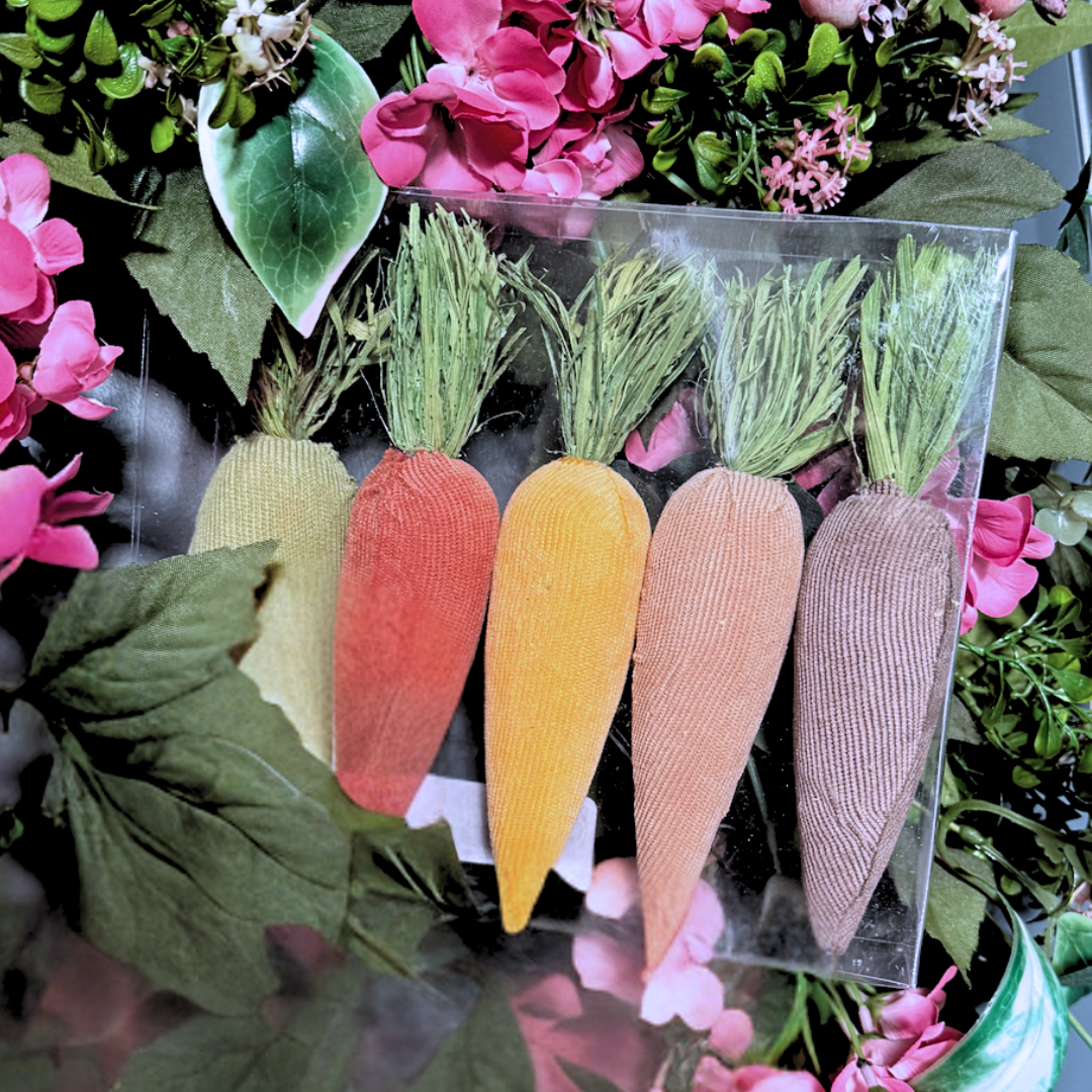 Set of colorful plush fabric carrots in a clear package with flowers and leaves in the background.
