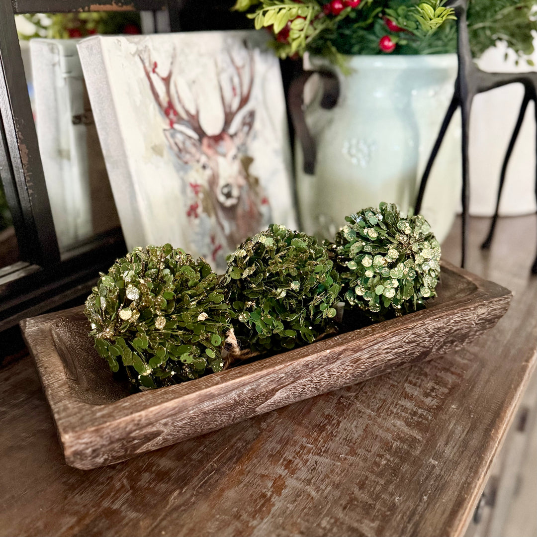 Decorative carved wooden dough bowl with gold glittered faux boxwood greenery ornaments on a wooden surface