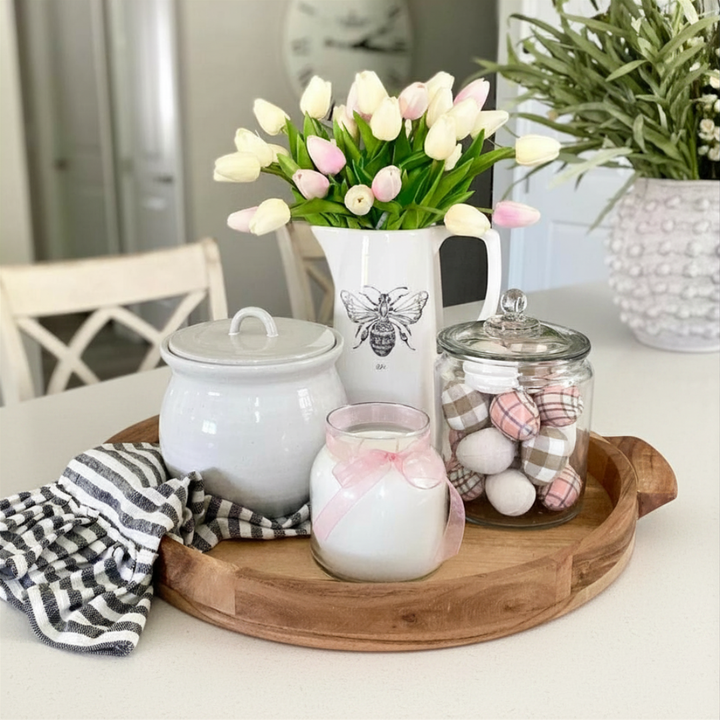 Decorative setting with a pitcher of tulips, jars, and a candle on a wooden tray.