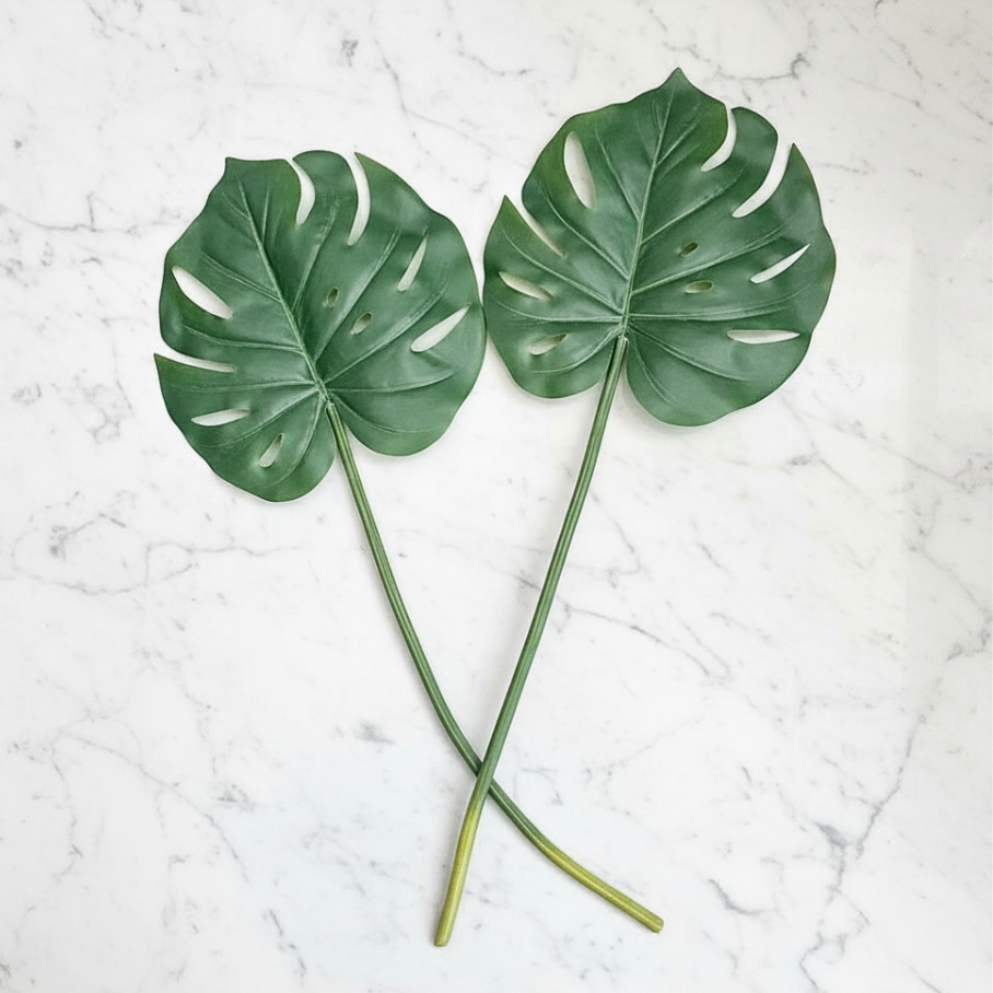 Two artificial green monstera leaves with long stems, arranged on a white marble surface.