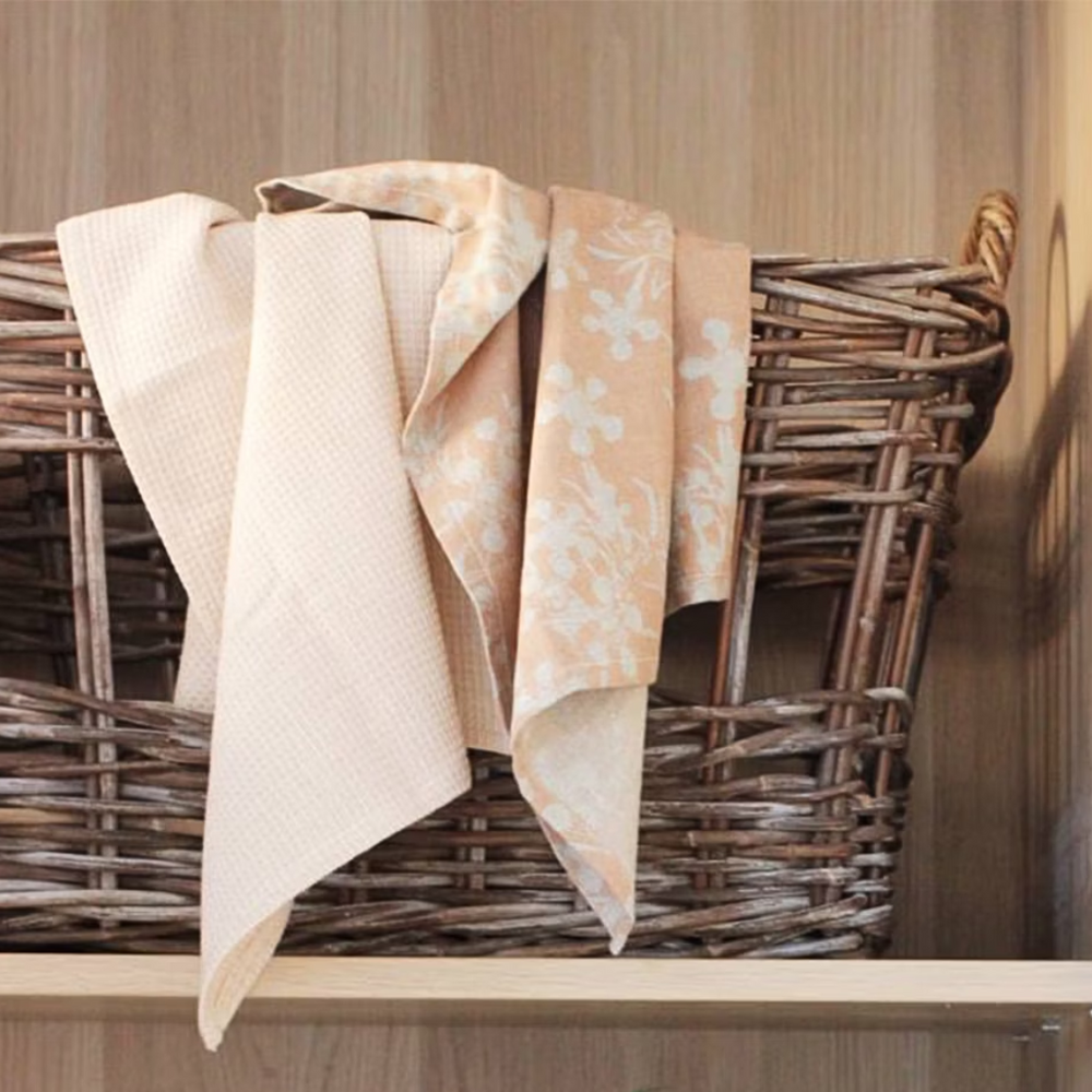 Two blush-colored cotton tea towels with a floral design on one and a honeycomb weave pattern on the other, draped over a wicker basket.