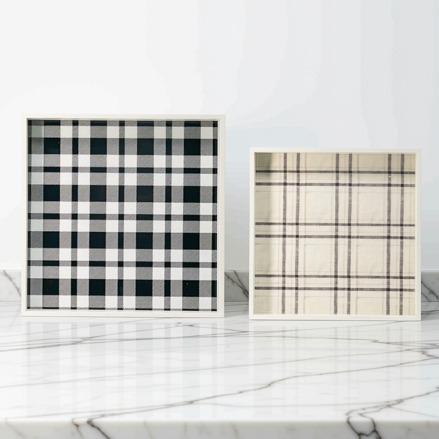 Two square framed plaid decorative  trays displayed on a white marble surface against a white wall. The left frame features a black and white buffalo plaid pattern, and the right frame features a beige and brown plaid pattern.