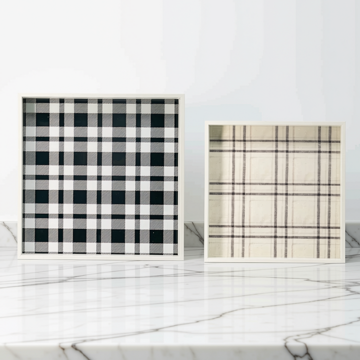 Two square framed plaid decorative  trays displayed on a white marble surface against a white wall. The left frame features a black and white buffalo plaid pattern, and the right frame features a beige and brown plaid pattern.