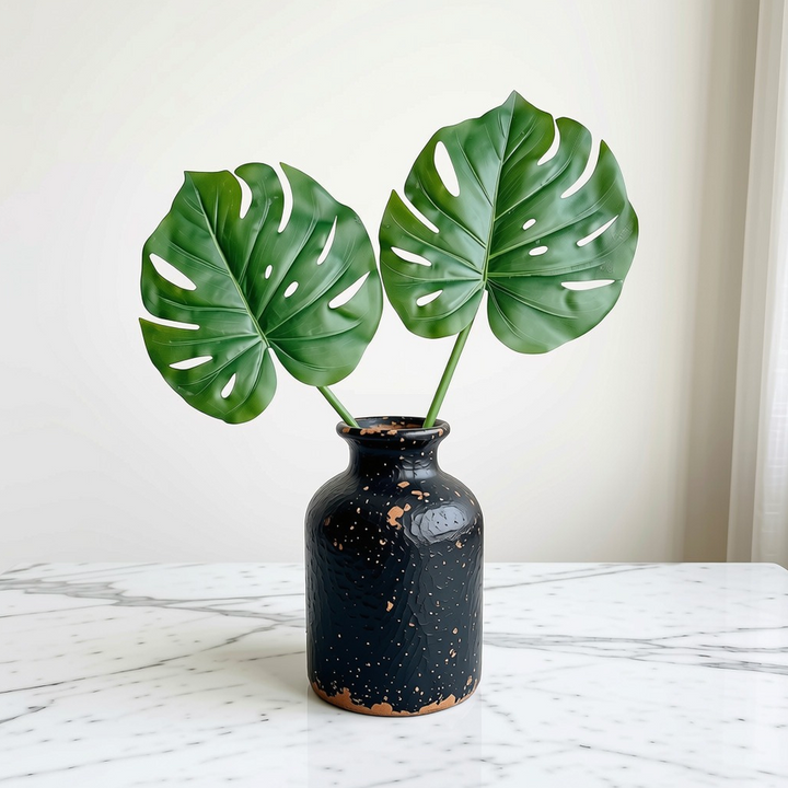 A black speckled ceramic vase with two green monstera leaves inserted, set on a white marble surface against a light beige wall.