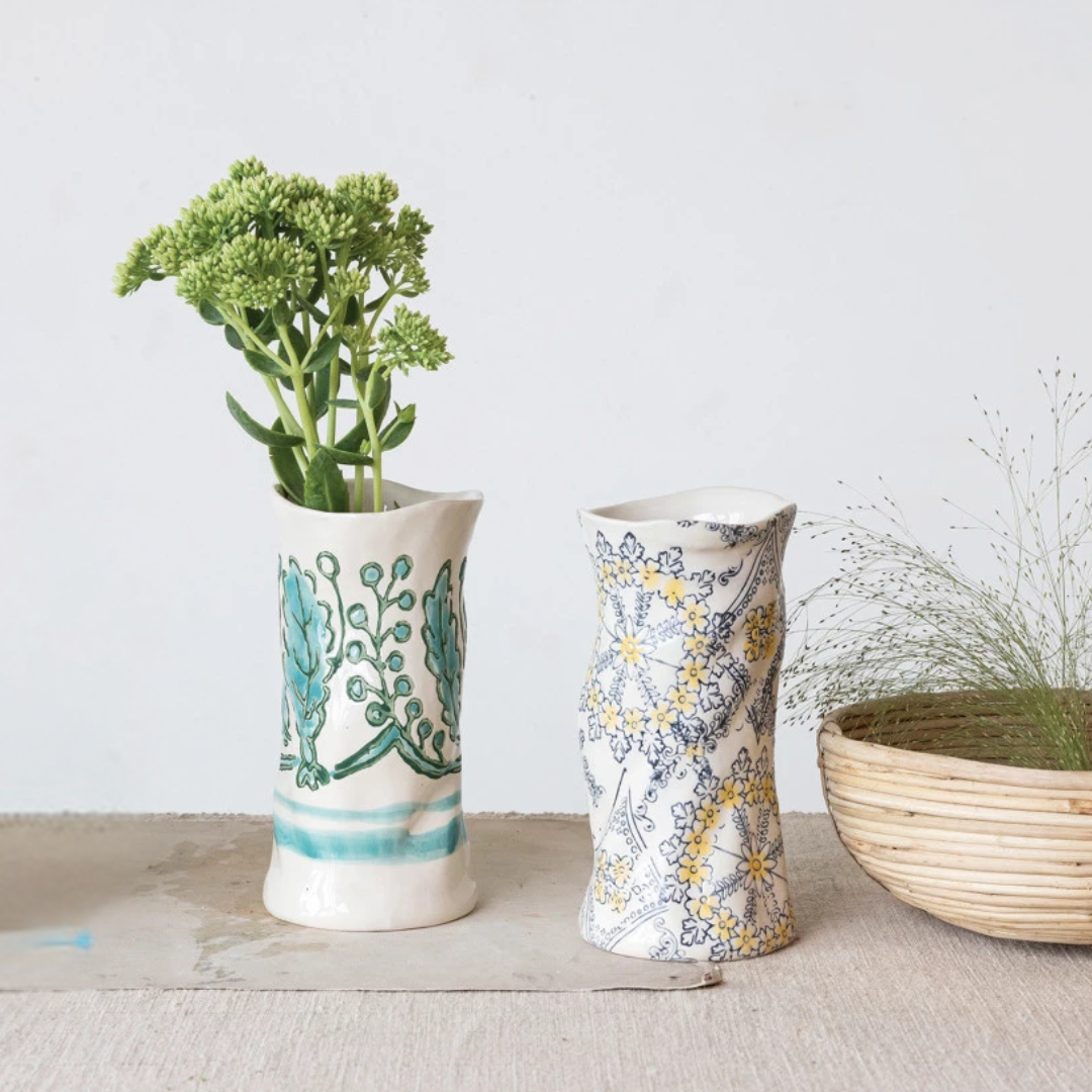 Two hand-painted stoneware vases, one with a blue and green floral pattern and the other with a beige and white floral pattern, displayed on a table with a basket in the background.