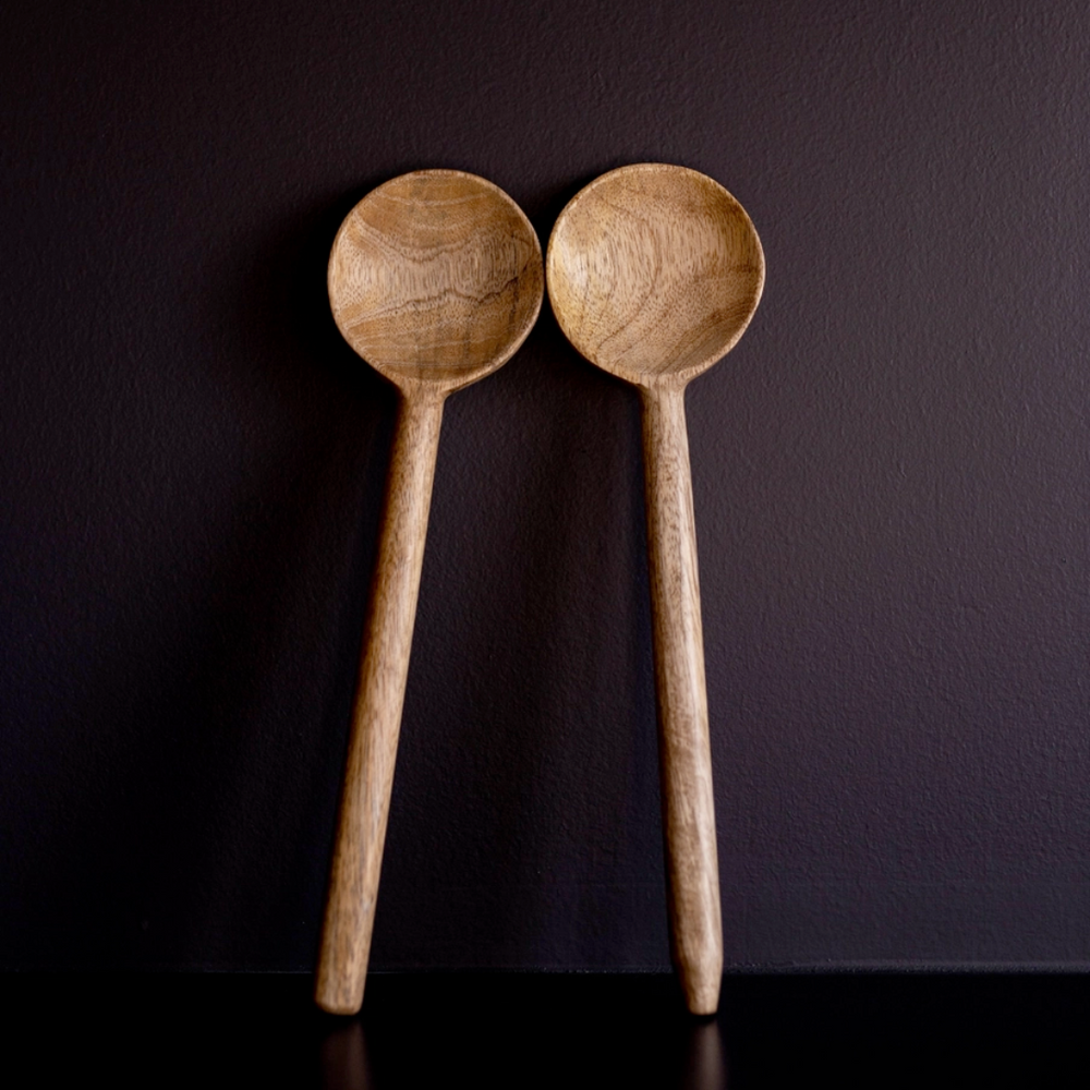 Two hand-carved mango wood serving spoons with a matte finish, standing upright against a dark background.