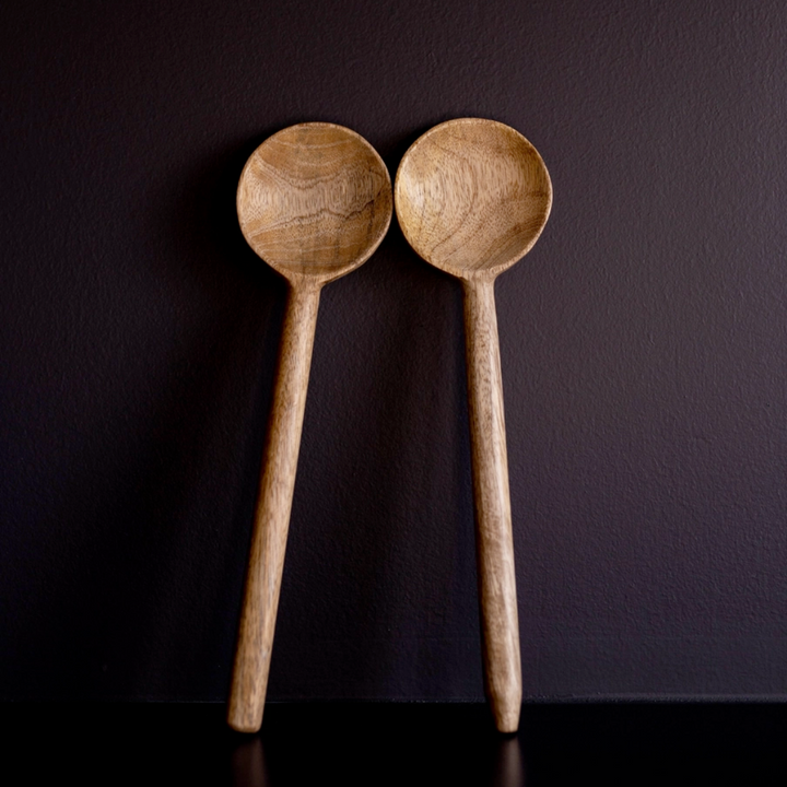 Two hand-carved mango wood serving spoons with a matte finish, standing upright against a dark background.