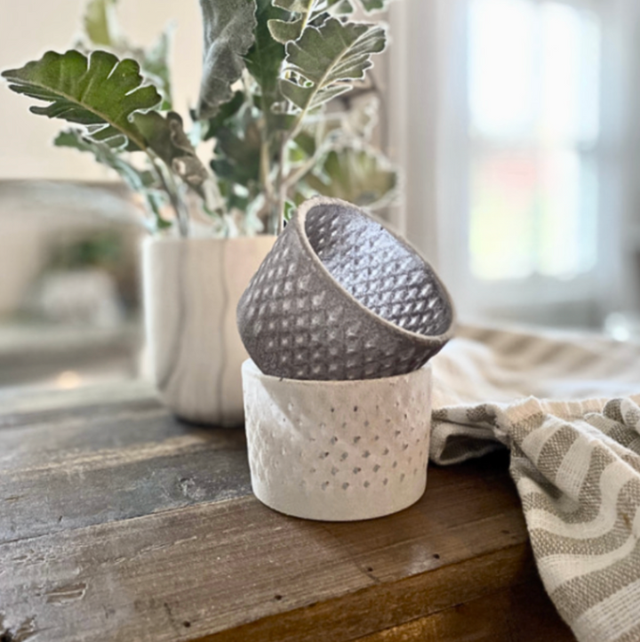 Two knobby glass votive candle holders, one in brownish/gray and one in white, displayed on a wooden surface with a plant in the background.