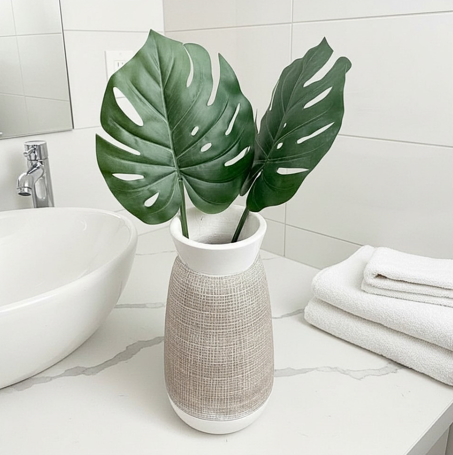 A white ceramic vase with a textured beige woven body, holding two large green monstera leaves, placed on a bathroom counter next to a white vessel sink and folded white towels.