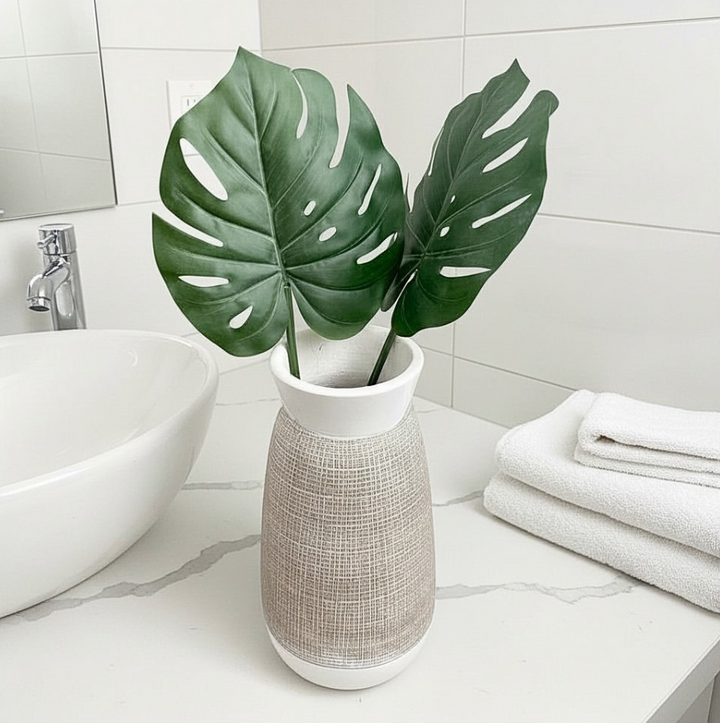A white ceramic vase with a textured beige woven body, holding two large green monstera leaves, placed on a bathroom counter next to a white vessel sink and folded white towels.