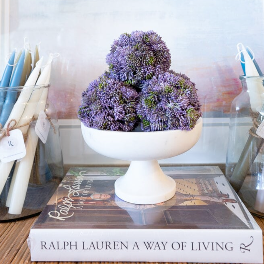 Decorative purple flower arrangement on a white pedestal over a Ralph Lauren book