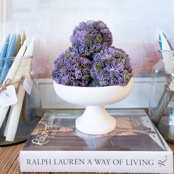Decorative purple flower arrangement on a white pedestal over a Ralph Lauren book