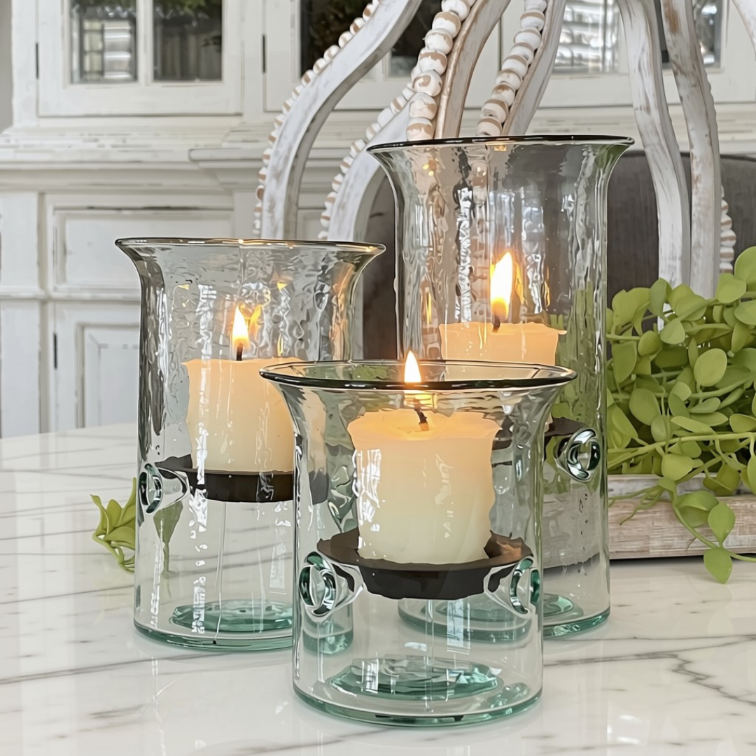 Three clear glass candle holders with a hammered finish, each containing a lit white pillar candle, arranged on a white marble surface with decorative greenery and a white chair in the background.
