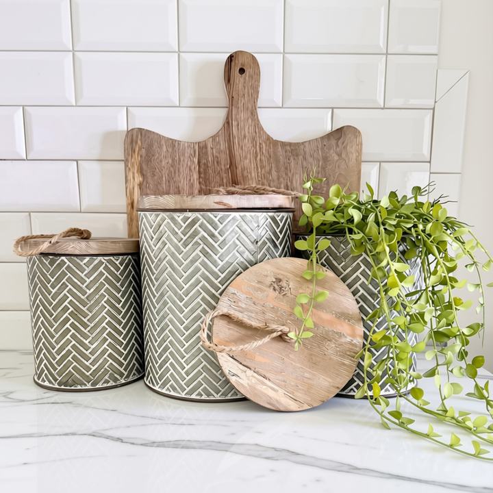 Three decorative metal canisters with a gray and white herringbone pattern and rustic wood lids, styled on a marble countertop with a wooden cutting board, plant, and white subway tile backsplash.