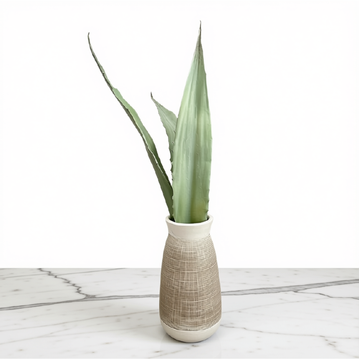 A tall green succulent or artificial plant stem displayed in a textured beige ceramic vase on a white marble surface against a white background.