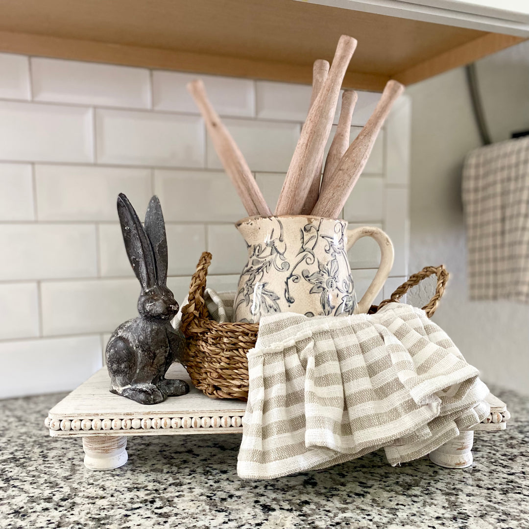 A set of three found wood roti pins displayed inside a home kitchen setting, with two pins leaning against a floral-patterned pitcher and a small metal rabbit figurine to the left.
