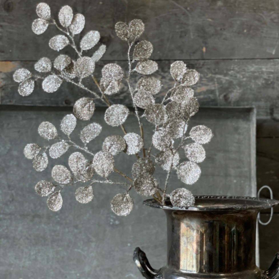 A sparkling eucalyptus spray with heavy glitter and mica finish, placed in a metal container, against a textured background.