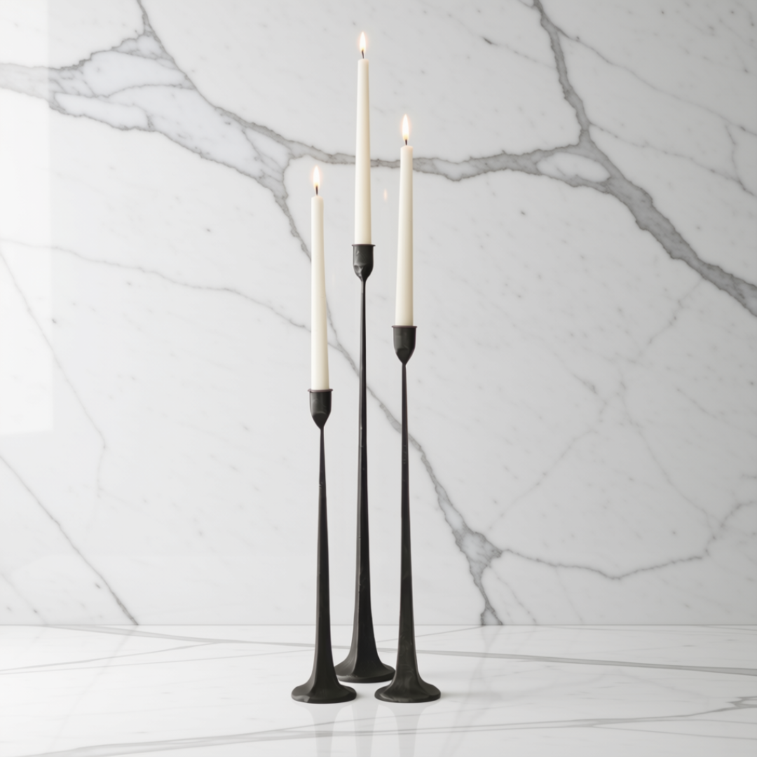 Three black cast iron taper candle holders with lit white taper candles, displayed on a white marble surface against a white marble wall background.