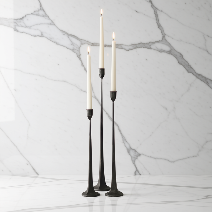 Three black cast iron taper candle holders with lit white taper candles, displayed on a white marble surface against a white marble wall background.