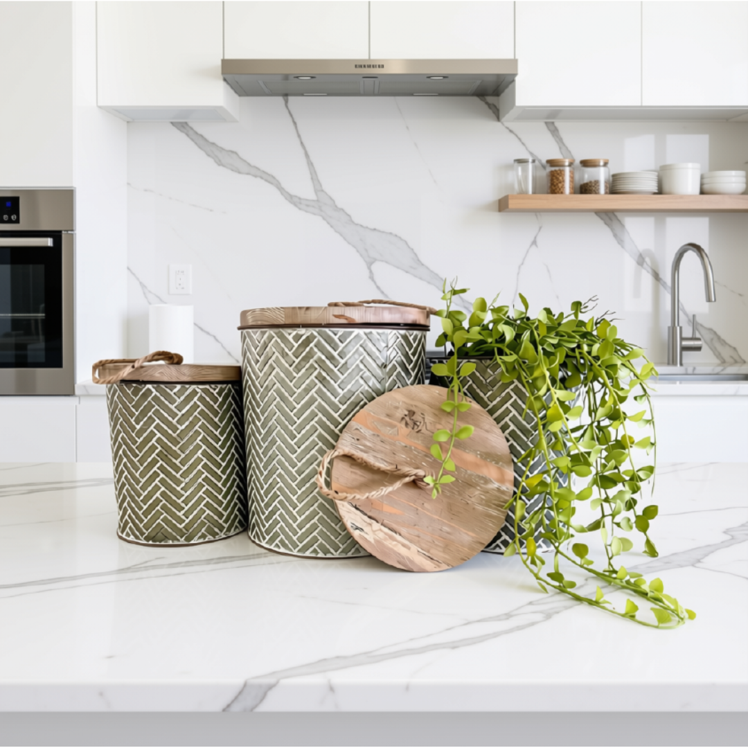 Three decorative metal canisters with a grey and white herringbone pattern and rustic wooden lids, styled on a white marble kitchen countertop next to a trailing green plant.