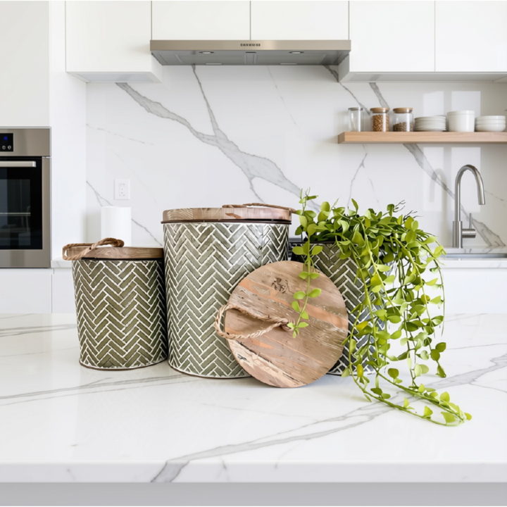 Three decorative metal canisters with a grey and white herringbone pattern and rustic wooden lids, styled on a white marble kitchen countertop next to a trailing green plant.