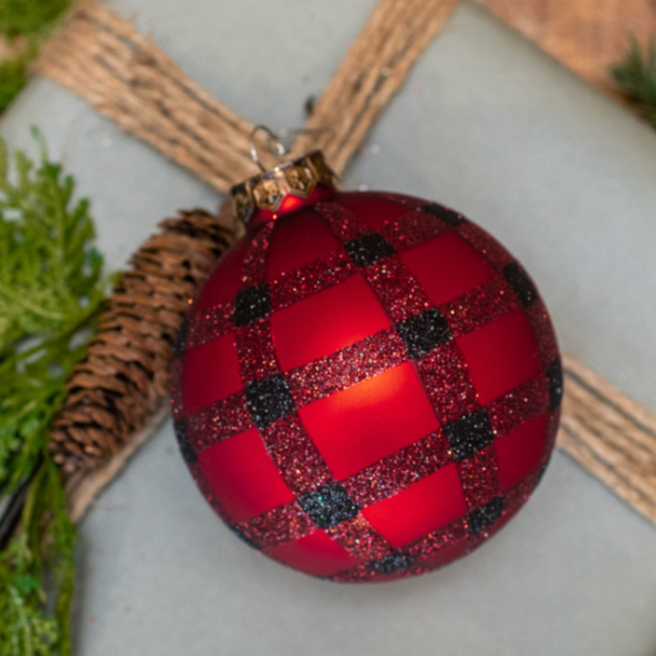 A red and black buffalo check patterned glass ornament on a Christmas tree, with a green and brown gift wrap and pine needles in the background.
