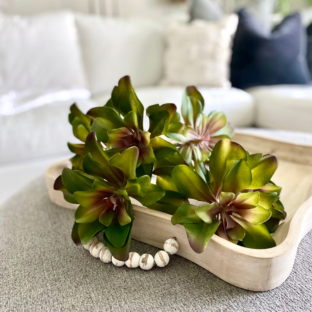 A faux succulent pick displayed in a wooden tray on a neutral colored surface, featuring green leaves with brown accents.