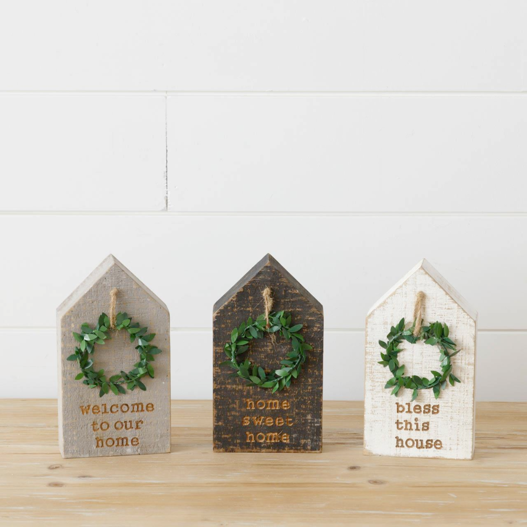Three wooden block houses with wreaths, each with a different color—beige, brown, and white—and each with a different phrase—'Welcome to our home', 'Home sweet home', and 'Bless this house'.