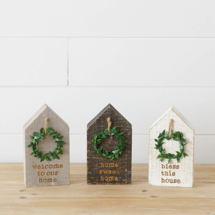 Three wooden block houses with wreaths, each with a different color—beige, brown, and white—and each with a different phrase—'Welcome to our home', 'Home sweet home', and 'Bless this house'.