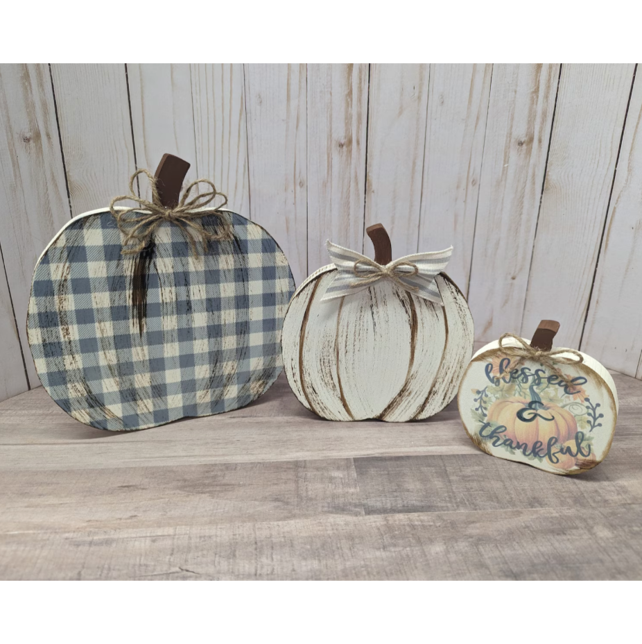 Three decorative autumn pumpkins displayed on a rustic wood surface against a whitewashed wood plank background. The left pumpkin is blue and white gingham plaid with a brown stem and bow.