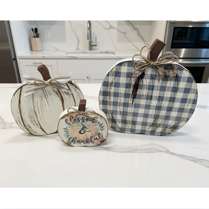 Three decorative autumn pumpkins displayed on a white marble countertop in a kitchen setting. The pumpkins include a distressed white pumpkin with a brown handle, a blue and white gingham pumpkin with a twine bow, and a small white pumpkin with a painted fall leaf and the words 'blessed & thankful'.