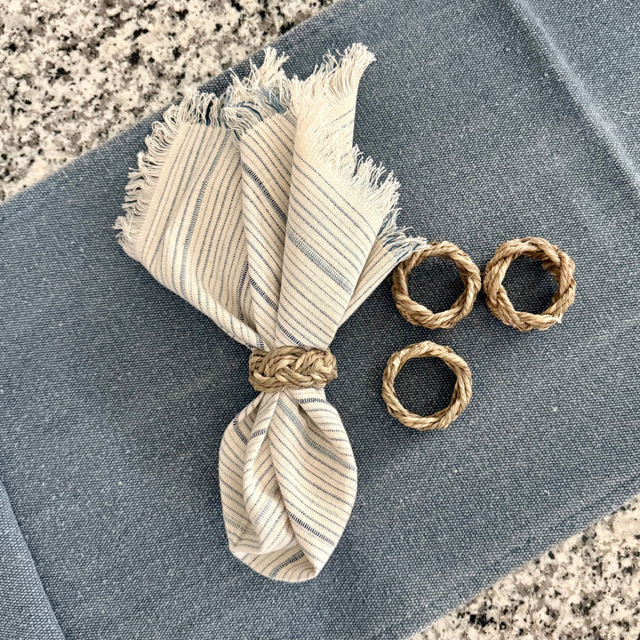 A set of four braided seagrass napkin rings displayed on a blue surface, alongside a frayed edge napkin in a matching earthy tone.