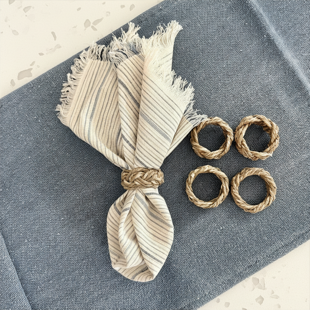 A set of four braided seagrass napkin rings displayed on a blue surface, alongside a frayed edge napkin in a matching earthy tone.
