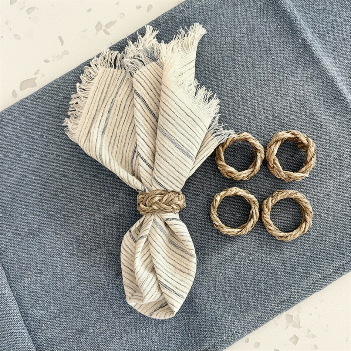 A set of four braided seagrass napkin rings displayed on a blue surface, alongside a frayed edge napkin in a matching earthy tone.